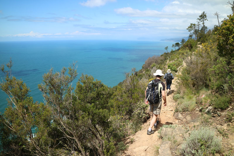 Two hikers walk along a narrow, dirt trail overlooking a vast blue ocean. Sparse vegetation surrounds them, and the landscape features rolling hills under a partly cloudy sky.